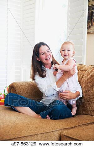 Stock Photo - Smiling mother holding smiling blonde infant girl while sitting on a couch.. Fotosearch