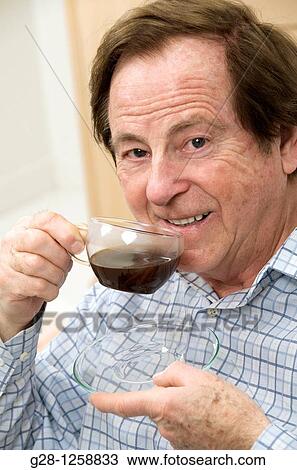 Stock Image - smiling senior man holding a cup of coffee in one hand and a saucer in the other hand about to drink coffee. Fotosearch