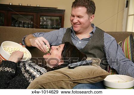 Snacking while lounging on the sofa at home is a fun pastime of otherwise busy couples Pictured here is a man feeding popcorn to his wife as she lays her head on his lap View Large Photo Image Stock Photo - Snacking while lounging on the sofa at home is a fun pastime of otherwise busy couples Pictured here is a man feeding popcorn to his wife as she lays her head on his lap. Fotosearch