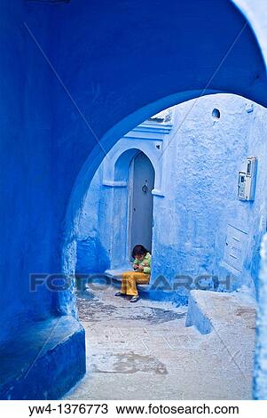 Stock Image - Street scene with a girl sitting on a threshold of a house, Chefchaouen, Morocco. Fotosearch