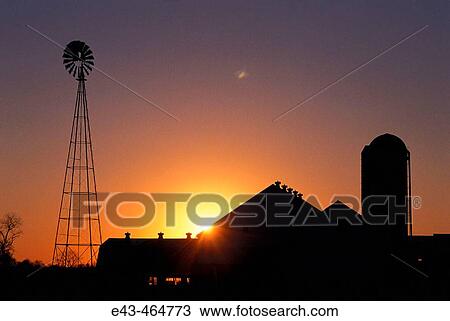 Stock Image - Sun rising over farm buildings with a windmill. Fotosearch