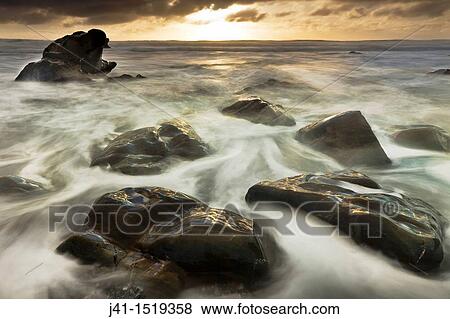 Stock Photo - Sun sets beneath heavy cloud layer, seascape on rocks off Gillespie's Beach, Westland National Park, West Coast, South Island, New Zealand. Fotosearch