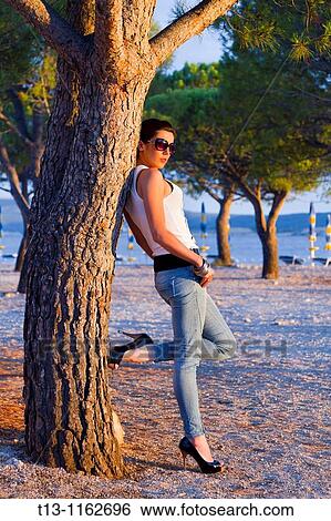 Stock Photograph - Sunset lit young woman inclined on a tree. Fotosearch