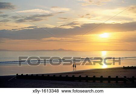 Sunset over the beach at the holiday seaside resort town of Barmouth in Gwynedd west Wales, UK Young couple strolling on beach View Large Photo Image Stock Photo - Sunset over the beach at the holiday seaside resort town of Barmouth in Gwynedd west Wales, UK Young couple strolling on beach. Fotosearch