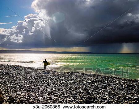 Surfer stood on a pebble beach looking out to sea with a rainstorm in the distance. Cornborough Range, Abbotsham, North Devon, England. View Large Photo Image Stock Photograph - Surfer stood on a pebble beach looking out to sea with a rainstorm in the distance. Cornborough Range, Abbotsham, North Devon, England.. Fotosearch
