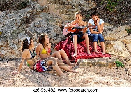 Stock Image - Teenage boy playing the guitar along with three girls at the beach. Fotosearch