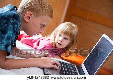 Stock Image - Teenage Boy with Kid Girl Lying Down and Using Laptop Computer. Fotosearch