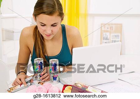 Stock Image - Teenage girl doing her homework and using her computer, while eating sweets, cookies, and drinking energy drinks. Fotosearch
