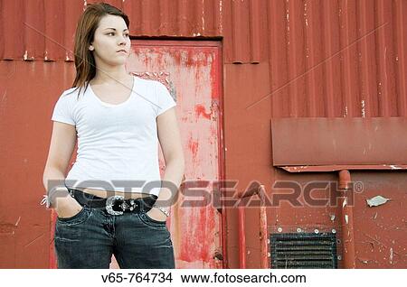 Picture - Teenage girl wearing white t_shirt standing in front of red corrugated iron building, looking moody. Fotosearch