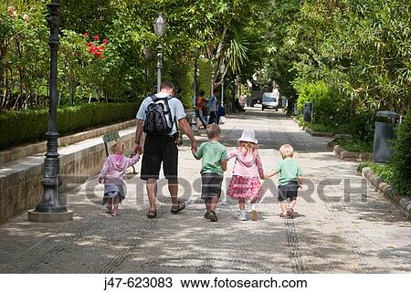 Stock Image - The back and legs of a man walking down a street holding four small childrens hands. Fotosearch