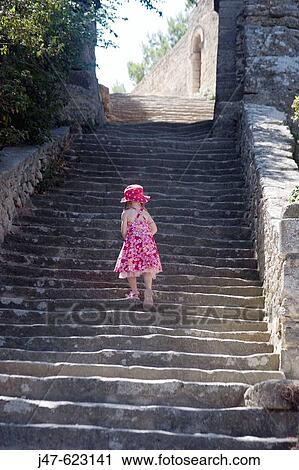 Stock Image - The back of a 5 year old girl in a red sundress, walking up a massive flight of ancient steps, in the sunshine. Fotosearch