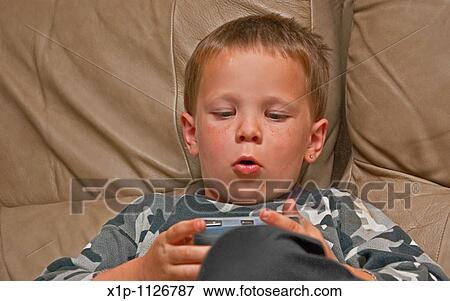Stock Photo - This 5 year old Caucasian boy with freckles is concentrating as he's playing an electronic video game on a beige colored leather couch indoors. Fotosearch