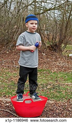 Stock Photograph - This Caucasian 5 year old boy is playing outside and standing on a red bucket that is frozen solid ice He's wearing a blue stocking cap, black sweat pants and holding a throwing toy Background intentionally blurred to emphasize subject. Fotosearch