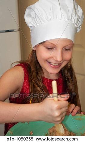 Stock Photography - This cute 9 year old Caucasian girl is happy and smiling as she's stiring a batch of cookie dough with a wooden spoon in a green bowl She's wearing a white chef's hat to complete the look. Fotosearch
