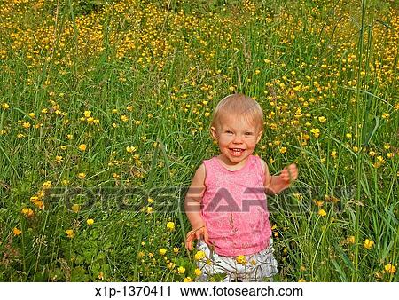 Stock Image - This cute Caucasian eighteen month old toddler girl is smiling and happy in long grass and buttercups She's wearing a pink shirt and has blue eyes and blond hair. Fotosearch
