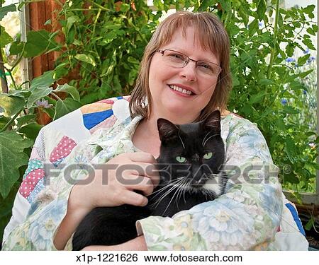 This middle aged woman is smilling and happy as she's holding a black cat with green eyes in a greenhouse She's wearing glasses and in a peaceful theme and setting View Large Photo Image Stock Photograph - This middle aged woman is smilling and happy as she's holding a black cat with green eyes in a greenhouse She's wearing glasses and in a peaceful theme and setting. Fotosearch