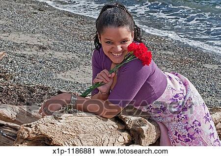 Stock Image - This pretty multi-racial woman in her thirties is smiling and happy on the beach She is holding some red carnations and laying on her stomach on some driftwood and wearing a floral pastel colored dress. Fotosearch