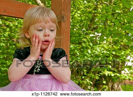 Stock Image - This stock image shows a two year old toddler girl outdoors in a pink princess dress with a surprised expression on her face Her little hands are on her cheeks with an? oh no? look Background intentionally blurred to emphasize subject. Fotosearch
