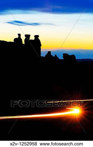 Three gossips at Arches National Park silhouetted against the sunset Car lights in the foreground trace the path of the road View Large Photo Image Stock Photo - Three gossips at Arches National Park silhouetted against the sunset Car lights in the foreground trace the path of the road. Fotosearch