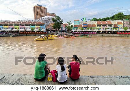 Stock Image - Three women sitting down on the Singapore River bank overlooking the brightly painted entertainment precinct of Clarke Quay, Singapore. Fotosearch