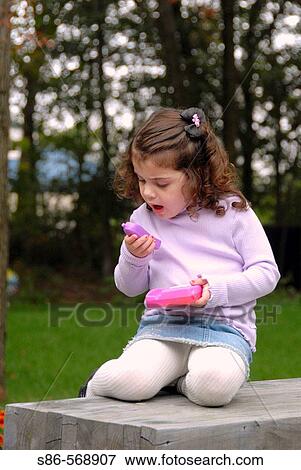 Stock Photo - Three year old girl, curled up on out door bench, talking into a toy telephone. Fotosearch