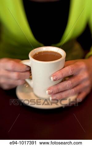 Stock Image - tight view of a woman's hands on a white coffee cup on a saucer on a table in a restaurant. Fotosearch
