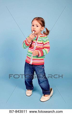 Stock Image - toddler, little girl, wearing grown up dress shoes, putting on lipstick, blue background, studio, Switzerland. Fotosearch