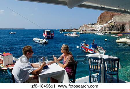 Stock Image - Tourist couple at restaurant at harbor with fishing boats at water with Oia above on mountain in Santorini Greece in Greek Islands. Fotosearch