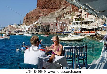 Tourist couple at restaurant at harbor with fishing boats at water with Oia above on mountain in Santorini Greece in Greek Islands View Large Photo Image Stock Photo - Tourist couple at restaurant at harbor with fishing boats at water with Oia above on mountain in Santorini Greece in Greek Islands. Fotosearch