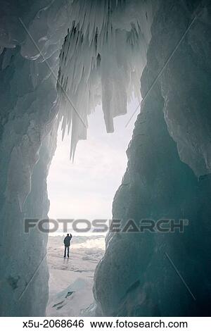 Stock Photograph - Tourist framed in the frozen mouth of the ice cave. Icicles formed on the roof of caves at Olkhon Island as the waves enter the cave and freeze at the beginning of winter.. Fotosearch