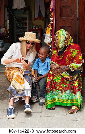 Tourist showing photos on her smart phone to local woman and child in East African market. View Large Photo Image Stock Image - Tourist showing photos on her smart phone to local woman and child in East African market.. Fotosearch