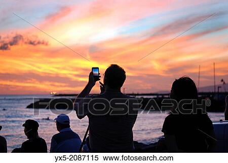 Stock Photography - Tourist taking picture of a spectacular sunset at Waikiki beach in Honolulu, Hawaii.. Fotosearch