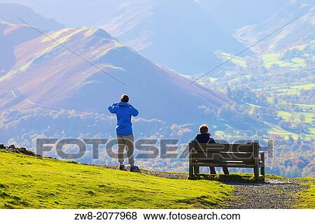 Stock Photo - Tourists standing and sitting on a bench, enjoying the view of Cat Bells in the Lake District National Park Cumbria England United Kingdom Great Britain.. Fotosearch