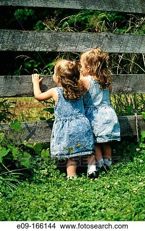 Twin toddler girls in denim dresses, looking between slats of wooden fence View Large Photo Image Picture - Twin toddler girls in denim dresses, looking between slats of wooden fence. Fotosearch