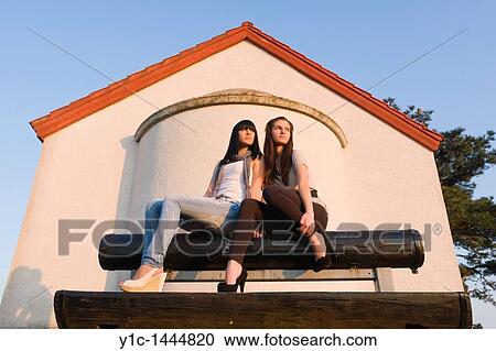 Stock Image - Two attractive young women are sitting on a wooden bench and watching sunset. Fotosearch