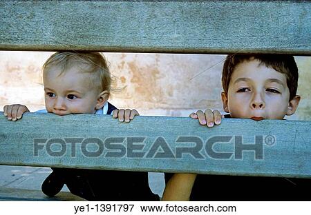 Two children making faces through the gaps in a park bench View Large Photo Image Stock Photo - Two children making faces through the gaps in a park bench. Fotosearch