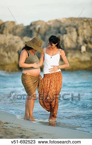 Stock Image - Two female friends, one woman is pregnant, walking along the beach. Fotosearch
