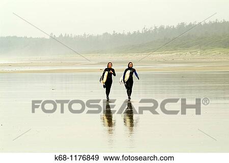 Stock Photo - two female surfers carry surfboards on the beach near Tofino, British Columbia, Canada. Fotosearch