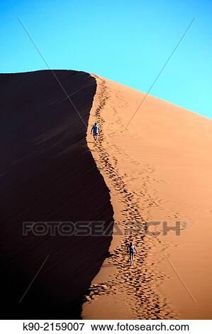 Stock Photo - Two people climbing up the sand dunes, sun with shadows, Sossusvlei, Namib-Naukluft Park, Namibia, Africa.. Fotosearch