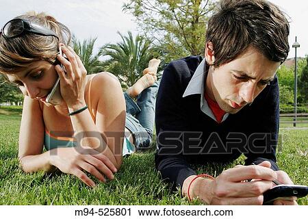 Stock Image - Two people on grass using palm pilot and cell phone. Fotosearch