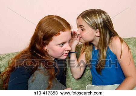 Stock Image - Two preteen girls sitting on a sofa, whispering to each other.. Fotosearch