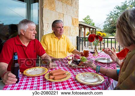 Stock Image - Two retired couples at a backyard barbecue party. Caucasian ethnicity, around 60 years old. Food and drinks, like fresh fruit, sausages, beer and wine, on the table.. Fotosearch