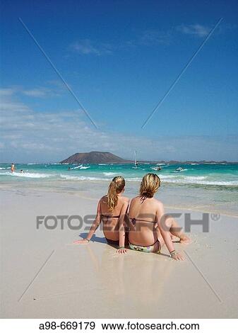 Stock Photo - two teenage girls sitting on sandy beach of Corralejo, Fuerteventura, Spain. Fotosearch