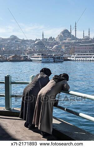 Stock Image - Two traditionaly dressed turkish women admire the view from the Galata bridge across the Golden Horn toward the Suleymaniye mosque and the Istanbul skyline. Istanbul, Turkey.. Fotosearch