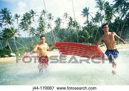 Stock Photo - Two young men running into the water with airbed at the beach. Fotosearch