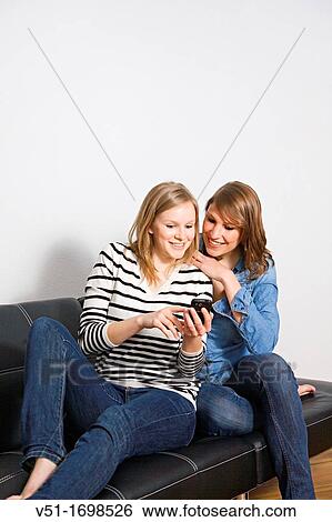 Two young women sitting on a couch sharing a sms View Large Photo Image Stock Photograph - Two young women sitting on a couch sharing a sms. Fotosearch