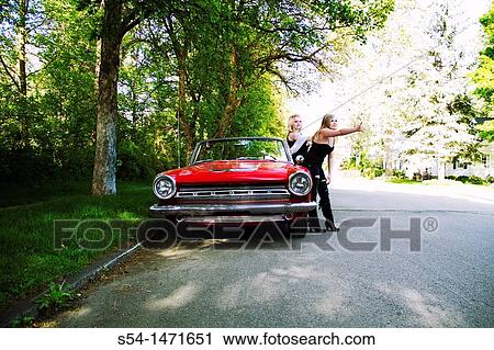 Stock Image - Two young women with a 1964 Dodge Dart in 1980s makeup, hitchhiking.. Fotosearch
