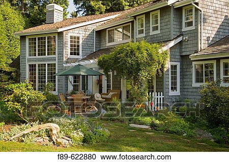 Stock Image - Umbrella over table & chairs on back_yard patio framed by vines, herbs, tulips (Tulipa cv.; Akebia quinata; Clematis cv.; Rosmarinus officinalis). Thompson, Blaine, WA.. Fotosearch