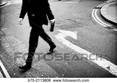 Unrecognizable Businessman crossing the street with a folder in hand and an arrow marking opposite direction painted on the asphalt. City of London, England, UK, Europe. View Large Photo Image Stock Photo - Unrecognizable Businessman crossing the street with a folder in hand and an arrow marking opposite direction painted on the asphalt. City of London, England, UK, Europe.. Fotosearch