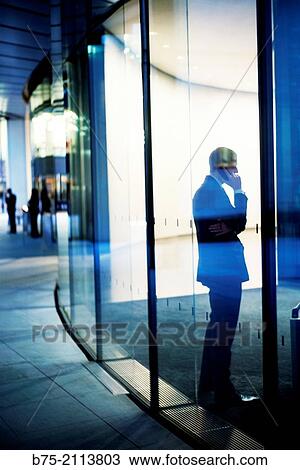 Stock Image - Unrecognizable Businessman talking on phone in an office corridor, seen through a glass in the City of London, England, UK, Europe.. Fotosearch
